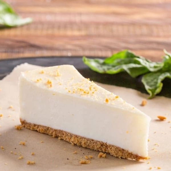 A slice of white cheesecake with a graham cracker crust, topped with crumbs, on parchment paper.