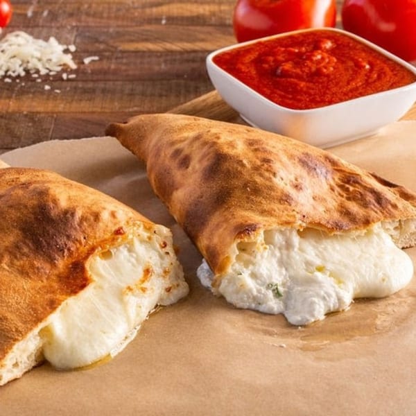 Two golden-brown calzones, one cut open to reveal melted cheese, with a side of marinara sauce.
